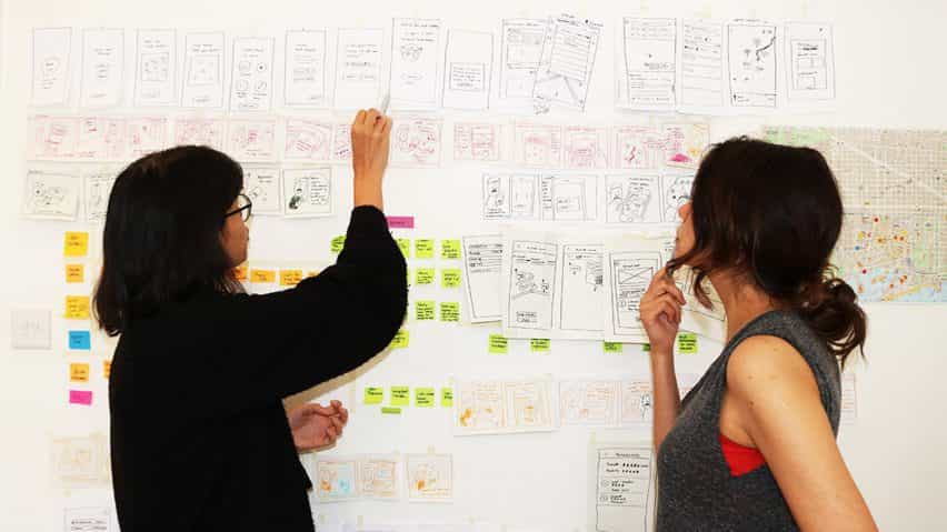 Dos personas de pie junto a una pared considerando los diagramas y notas adjuntas a ella