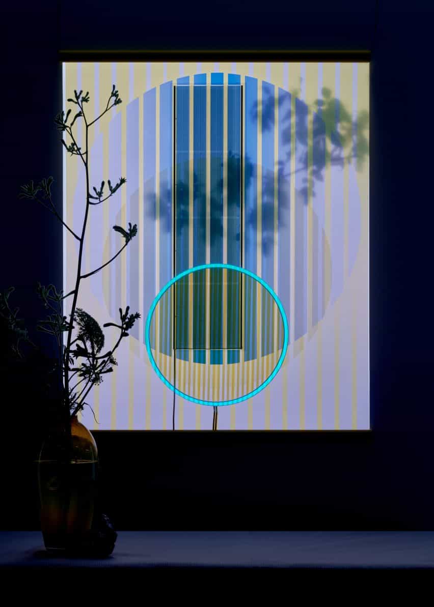 Tapiz solar de Marjan van Aubel fotografiado por la noche colgado detrás de un jarrón con flores con un anillo azul brillante en su centro